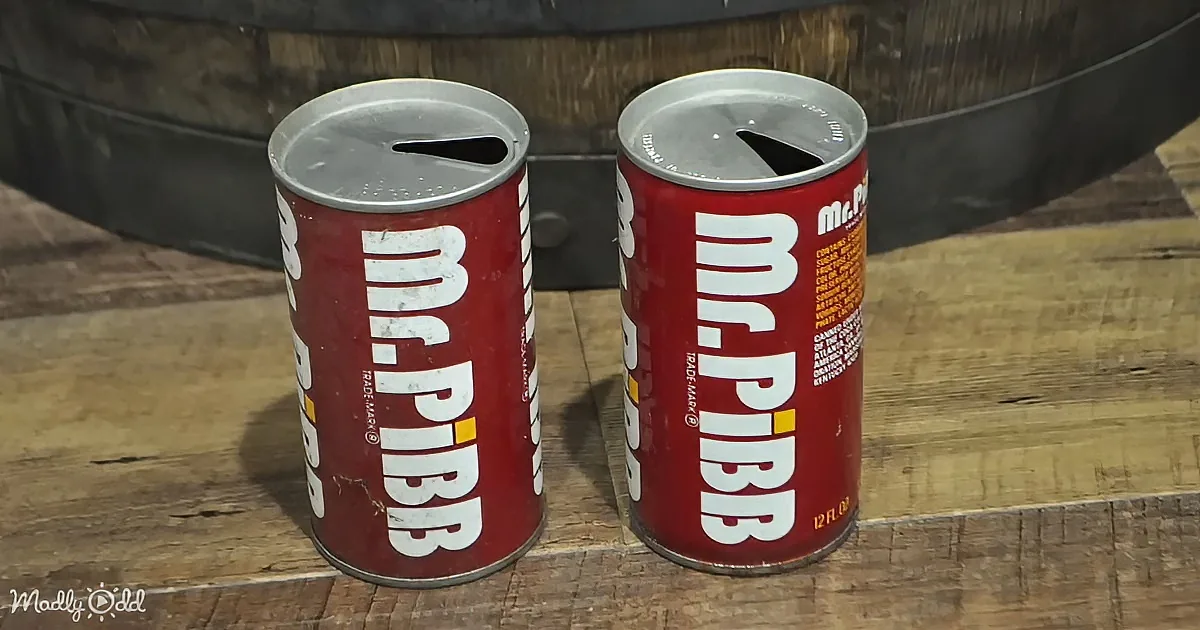 Two Mr. Pibb soda cans with holes in the tops sit on a wooden surface.