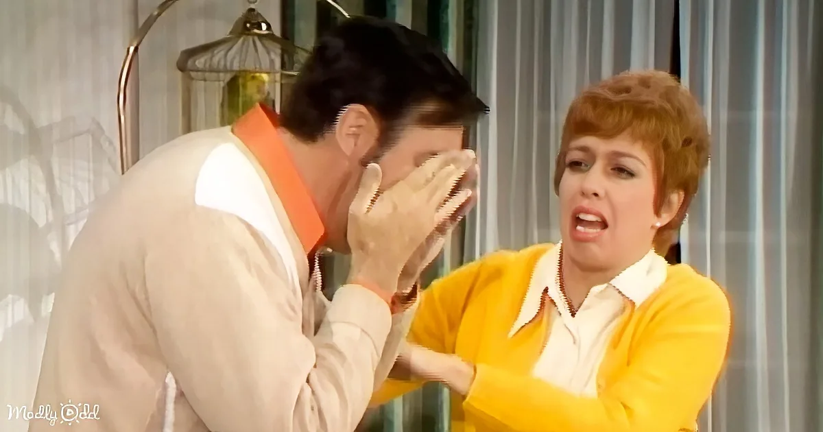 A man in a beige suit and orange shirt covers his face, while a woman in a yellow cardigan and white shirt looks concerned.