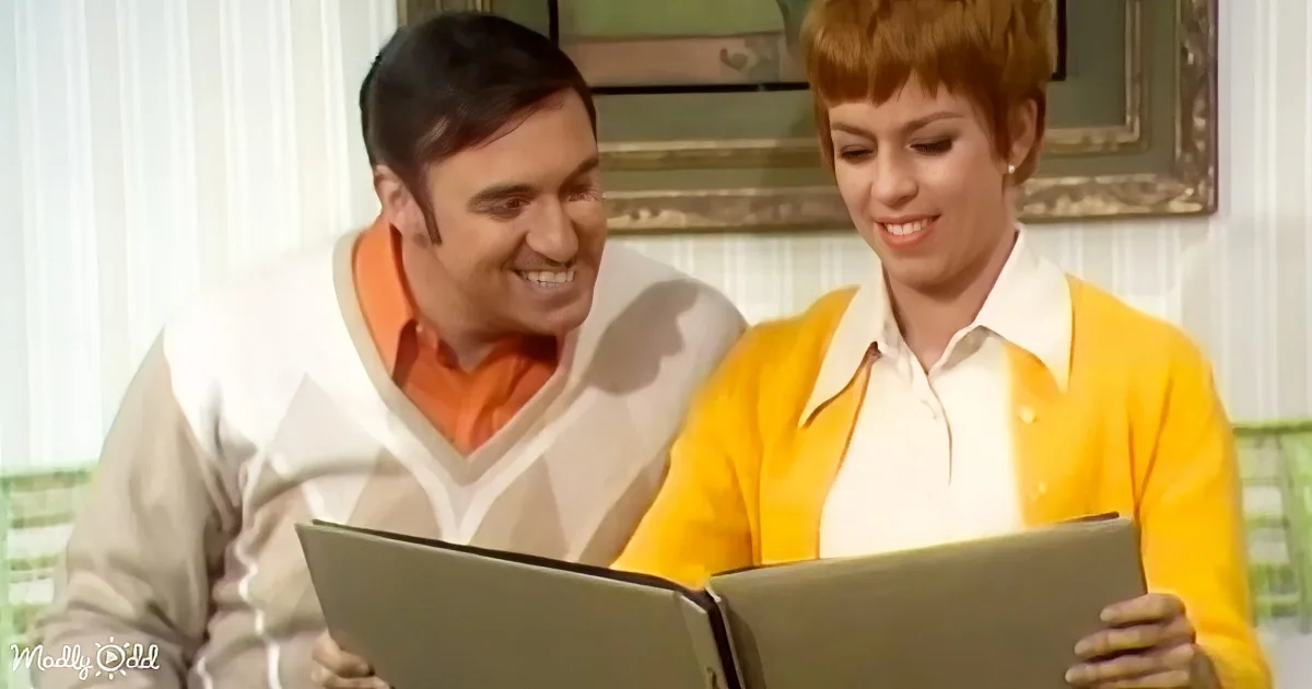 A man and woman in retro attire smile while looking at a book together.