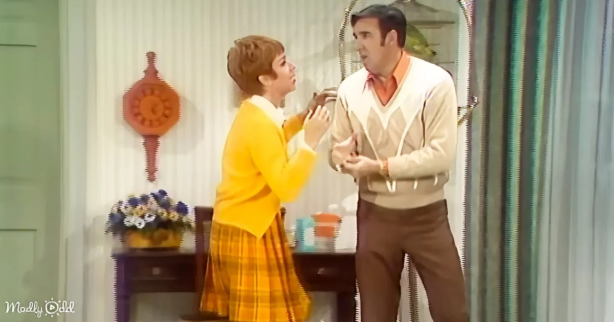 A woman in a yellow dress and man in a beige sweater are engaged in a conversation in a room with a clock and flowers.