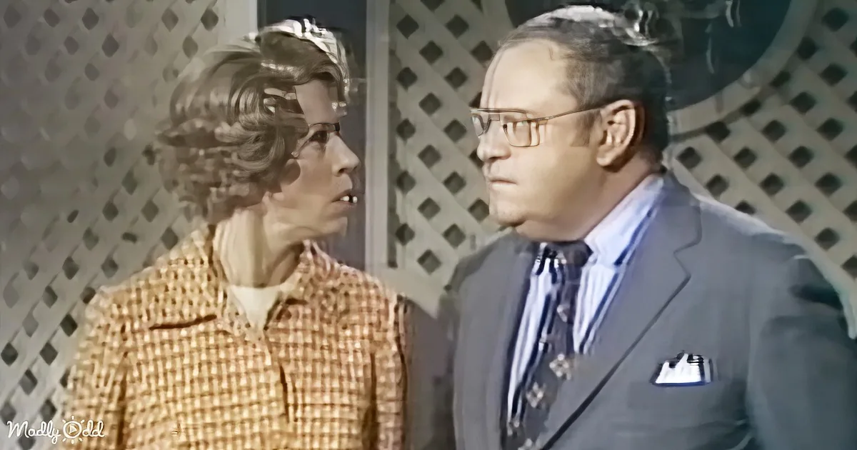 A man in a suit and tie stands next to a woman in a checkered jacket, both looking at each other.