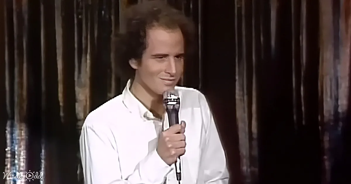 A man in a white shirt holds a microphone, smiling, against a dark curtain backdrop.
