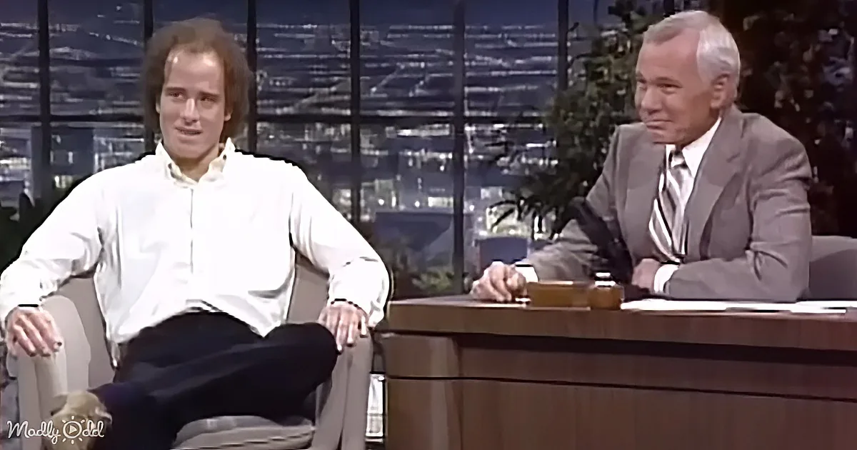 A man in a white shirt sits casually in a chair, while another in a suit sits behind a desk, both on a talk show set.