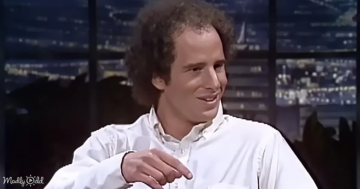 A man in a white shirt is smiling and gesturing with his hand, sitting in front of a cityscape backdrop.
