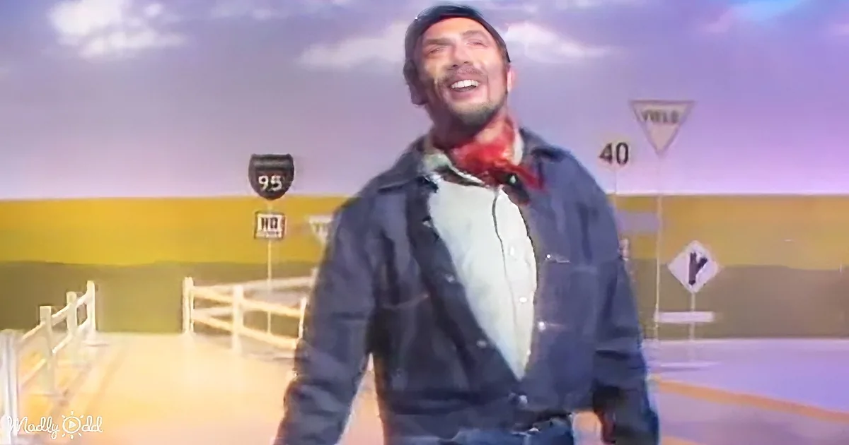 A man in a denim jacket and red bandana smiles in front of a road sign backdrop.