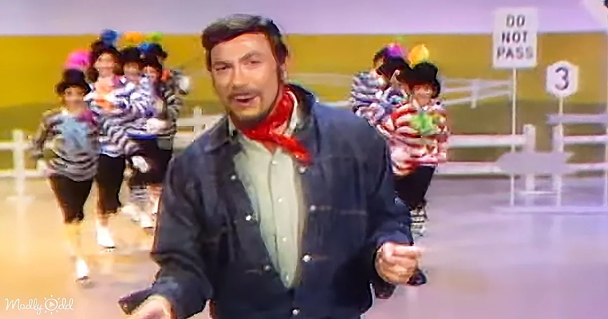 A man in a denim jacket and red scarf gestures while children in colorful outfits run behind him.