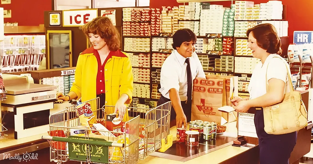 A cashier in a yellow jacket smiles as a customer hands over items at a vintage grocery store checkout.