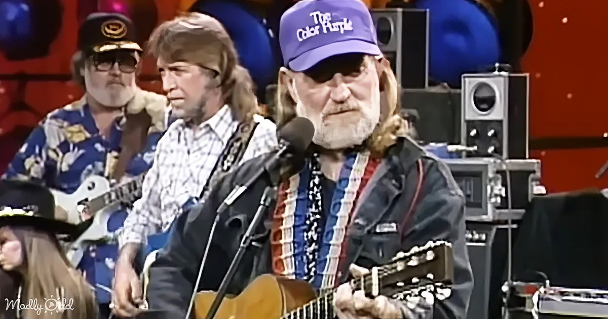 A man with a beard and a blue "The Color Purple" hat plays guitar and sings into a microphone.