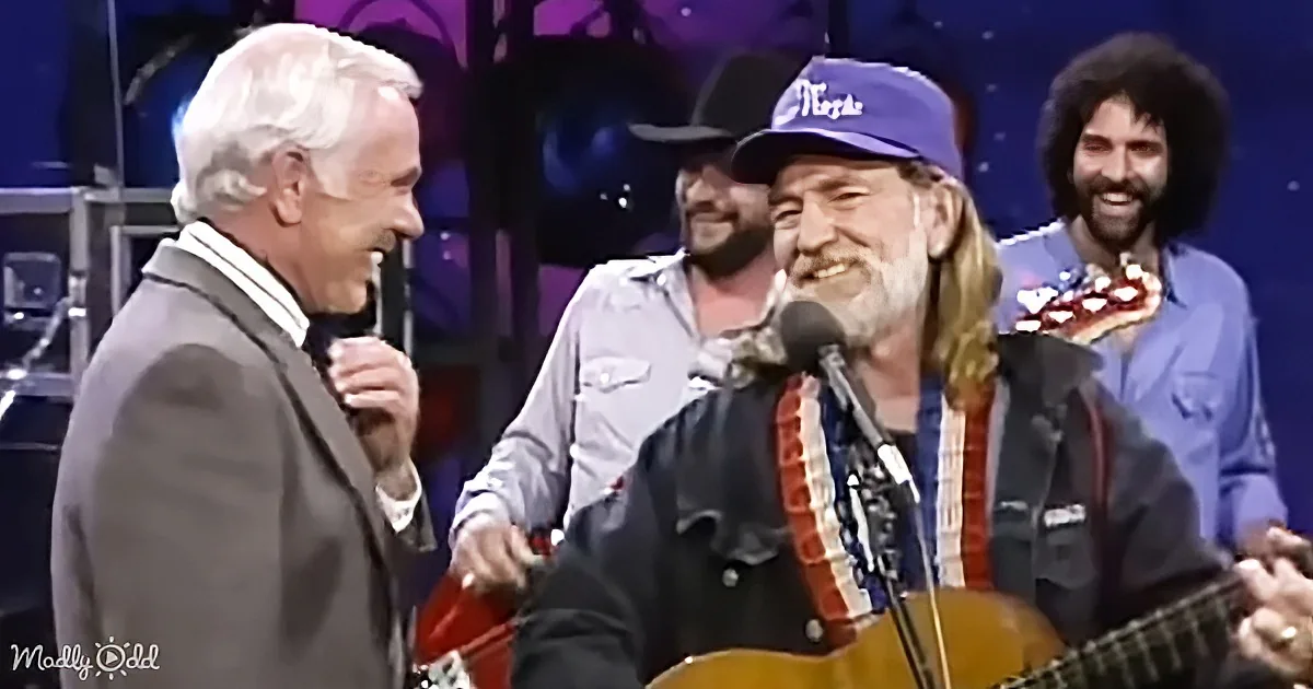 A man with a beard and hat plays guitar while another man in a suit looks on, both smiling.