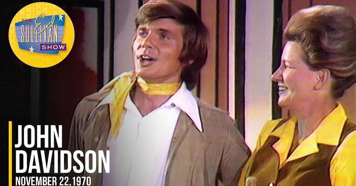 A man in a brown jacket and white shirt stands next to a woman in a yellow outfit, both looking up.