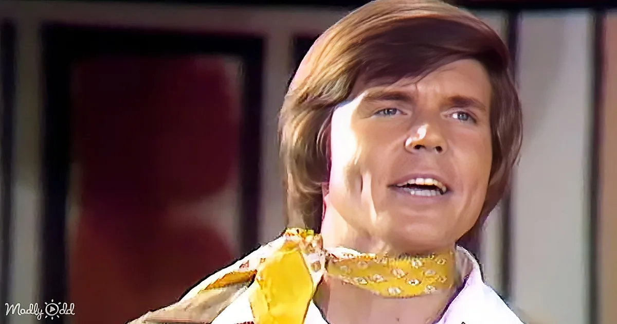 A person with shoulder-length brown hair, wearing a yellow patterned scarf and a white shirt, appears to be speaking or...