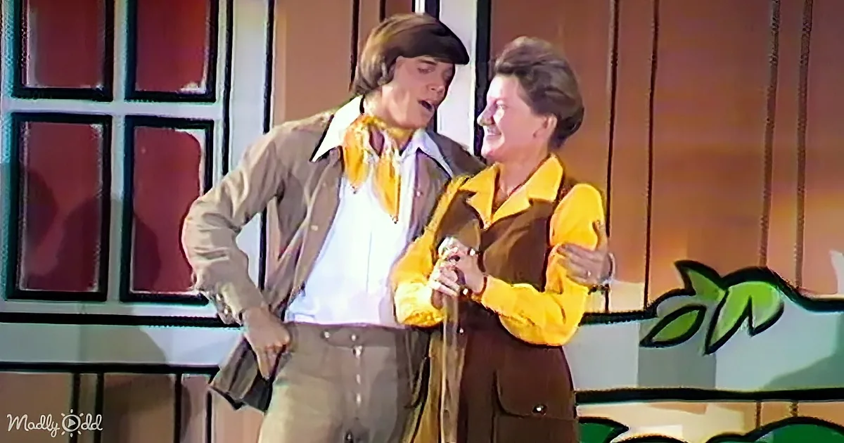 Two individuals in vintage attire, one in a brown jacket and the other in a yellow shirt, share a moment on stage.