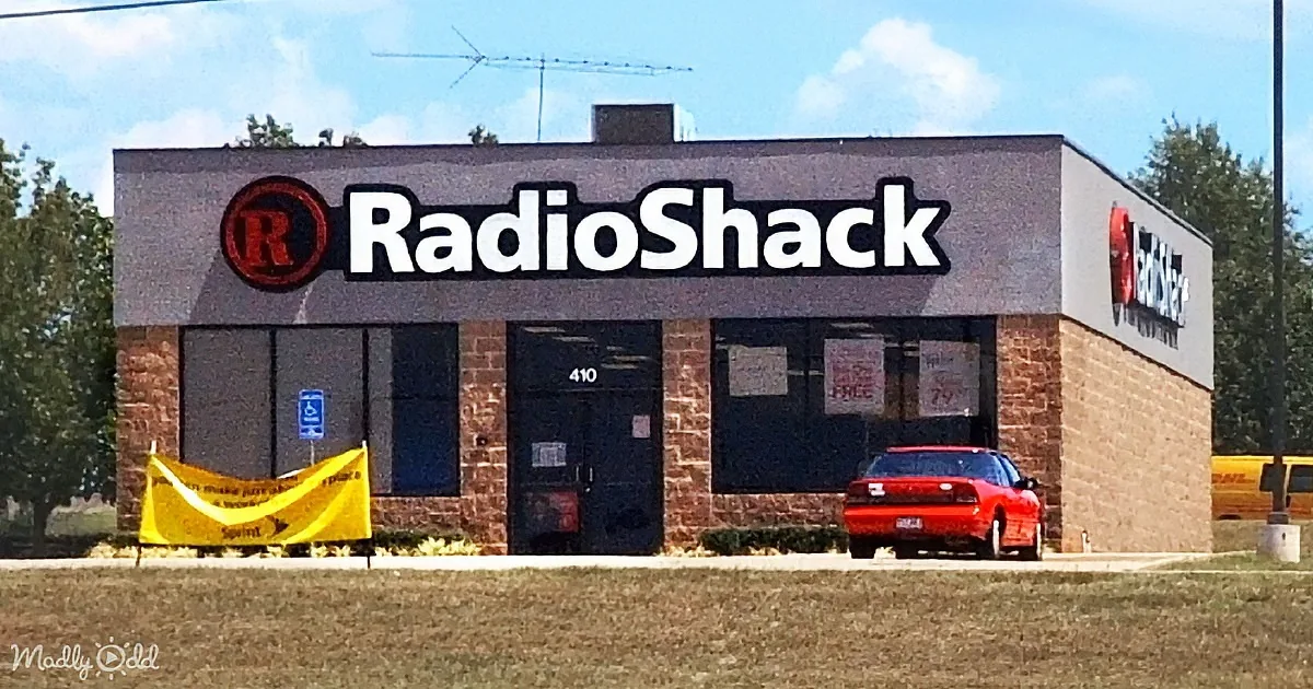 A RadioShack store with a red car parked in front and a yellow banner on the left.