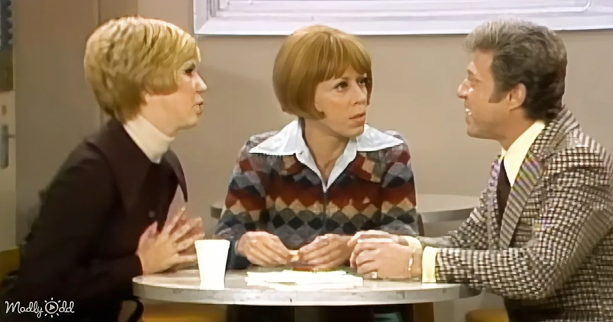 Three individuals are seated around a table, engaged in a discussion, with one person gesturing while others listen...