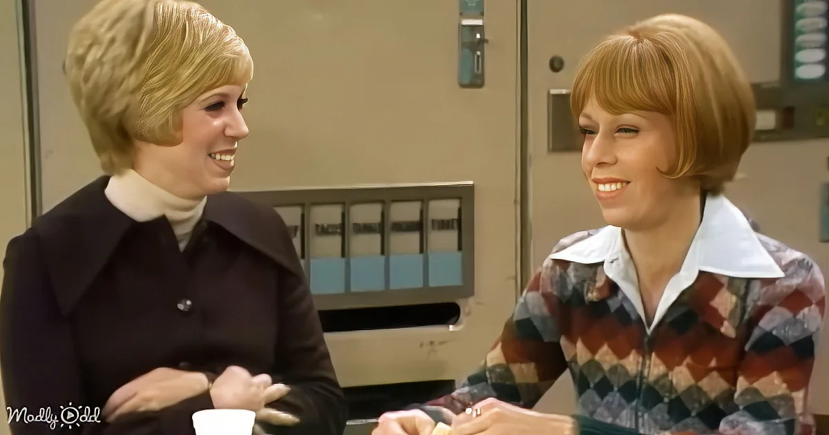 Two women in vintage attire smile and converse in a retro office setting.