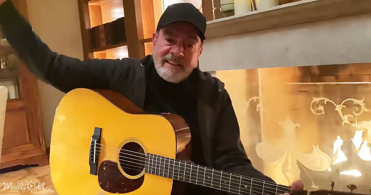 A man in a black hoodie and cap plays a yellow acoustic guitar in front of a lit fireplace.