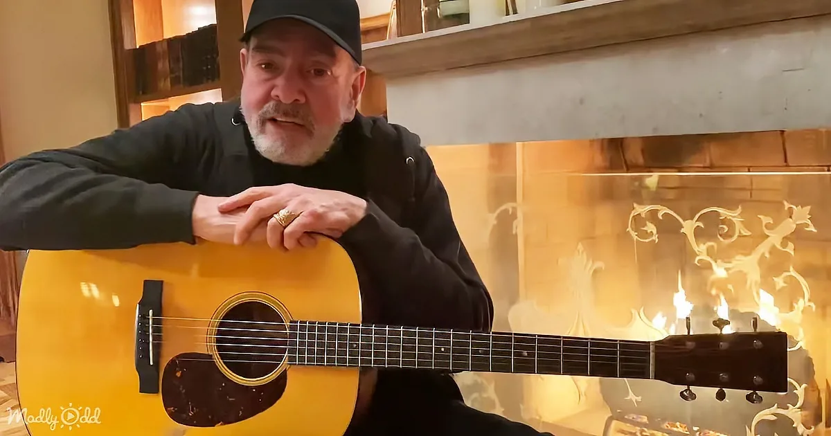 A man in a black shirt and cap plays an acoustic guitar in front of a lit fireplace.