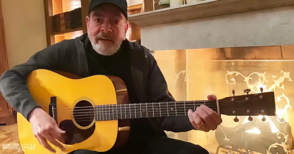 A man plays a yellow acoustic guitar in front of a lit fireplace.