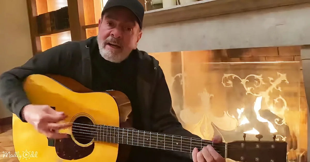 A man plays a yellow acoustic guitar in front of a lit fireplace.