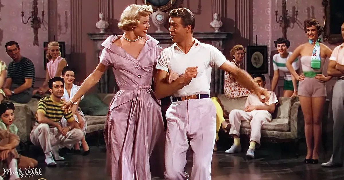 A couple in a pink dress and white shirt dances joyfully, surrounded by a lively crowd in a vintage setting.