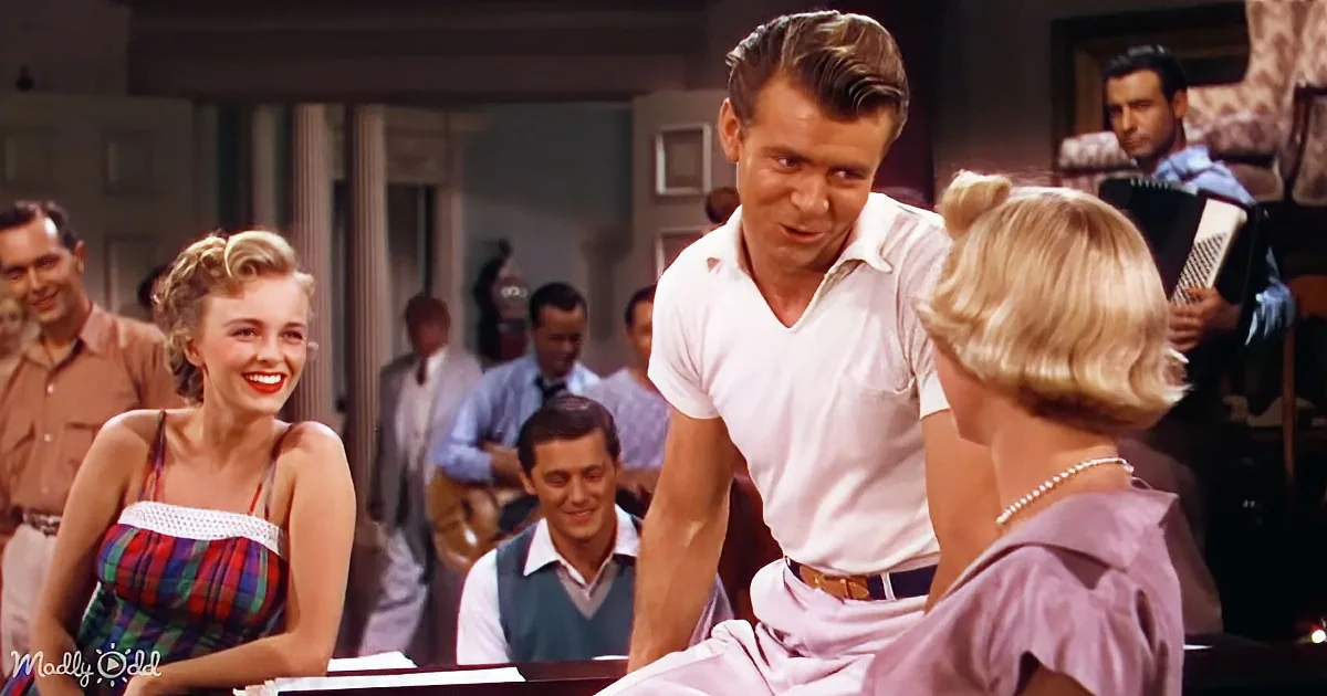 A man in a white shirt leans forward, engaging with a woman in a plaid dress at a piano, surrounded by other people in a...