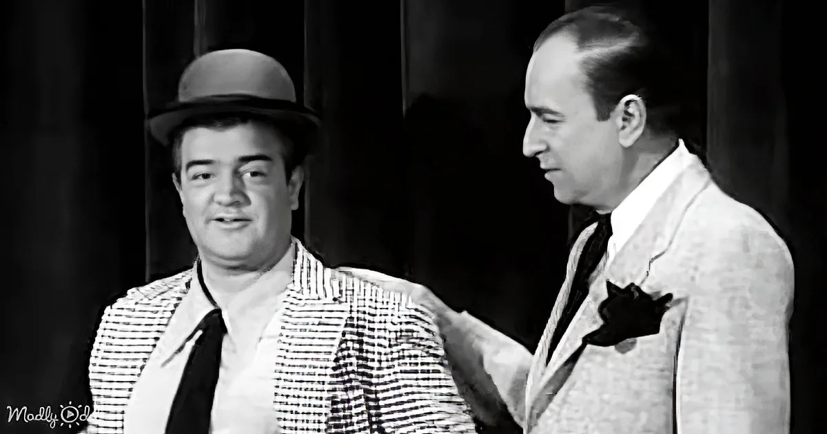 A man in a checkered shirt and bowler hat is being comforted by another man in a suit with a pocket square.