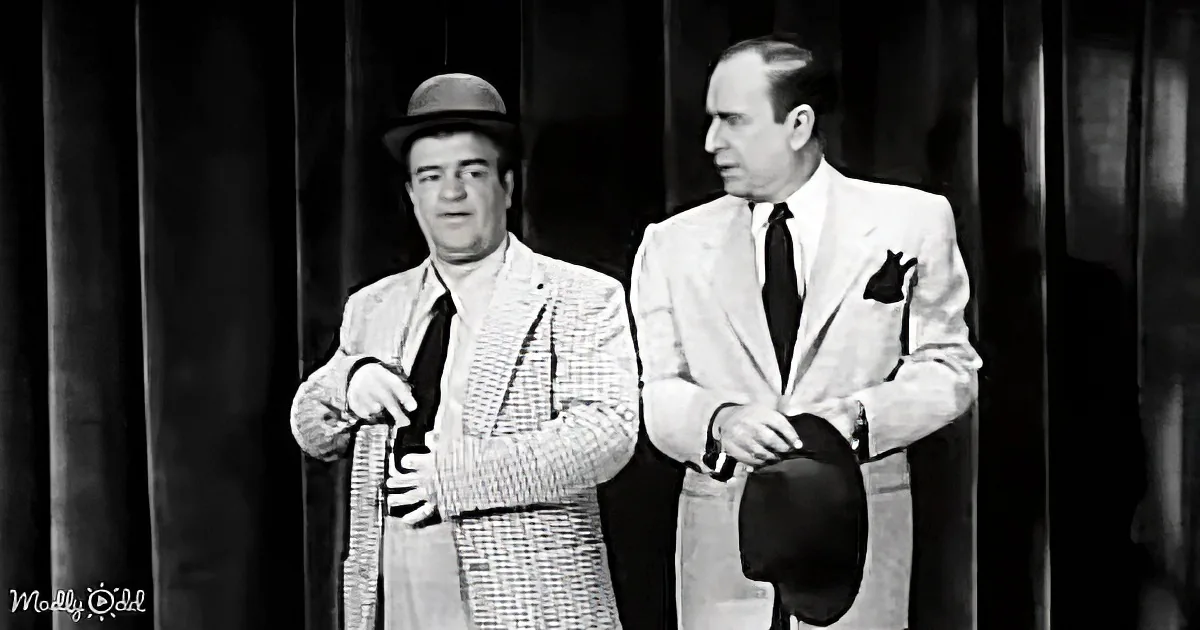Two men in suits stand side by side, one holding a cane, against a dark curtain backdrop.
