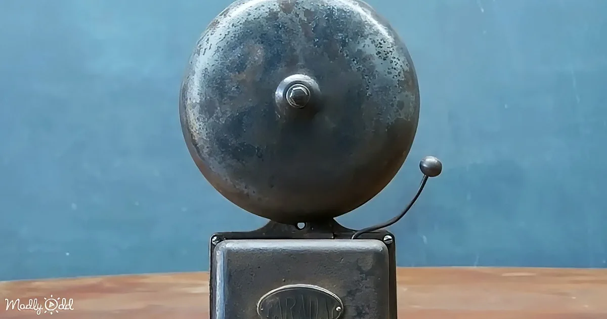 An old, weathered metal bell with a pull cord sits atop a rectangular base on a wooden surface.