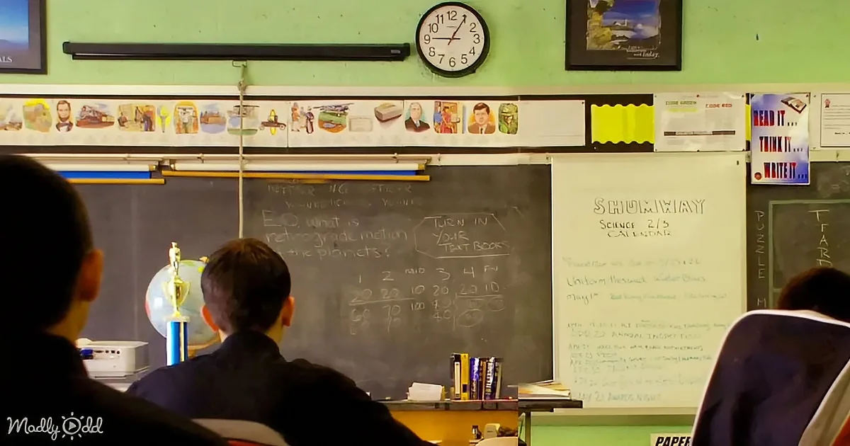 Students sit in a classroom with a chalkboard displaying science questions and a trophy on the desk.