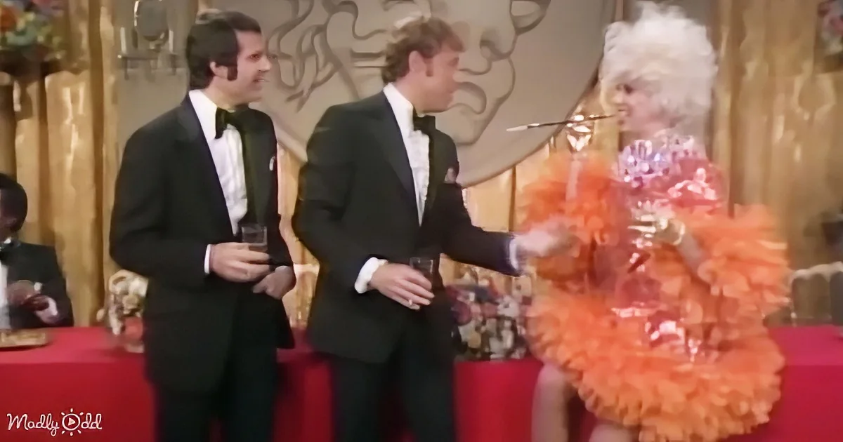 A man in a tuxedo gestures towards a person in a vibrant orange outfit with feathers.