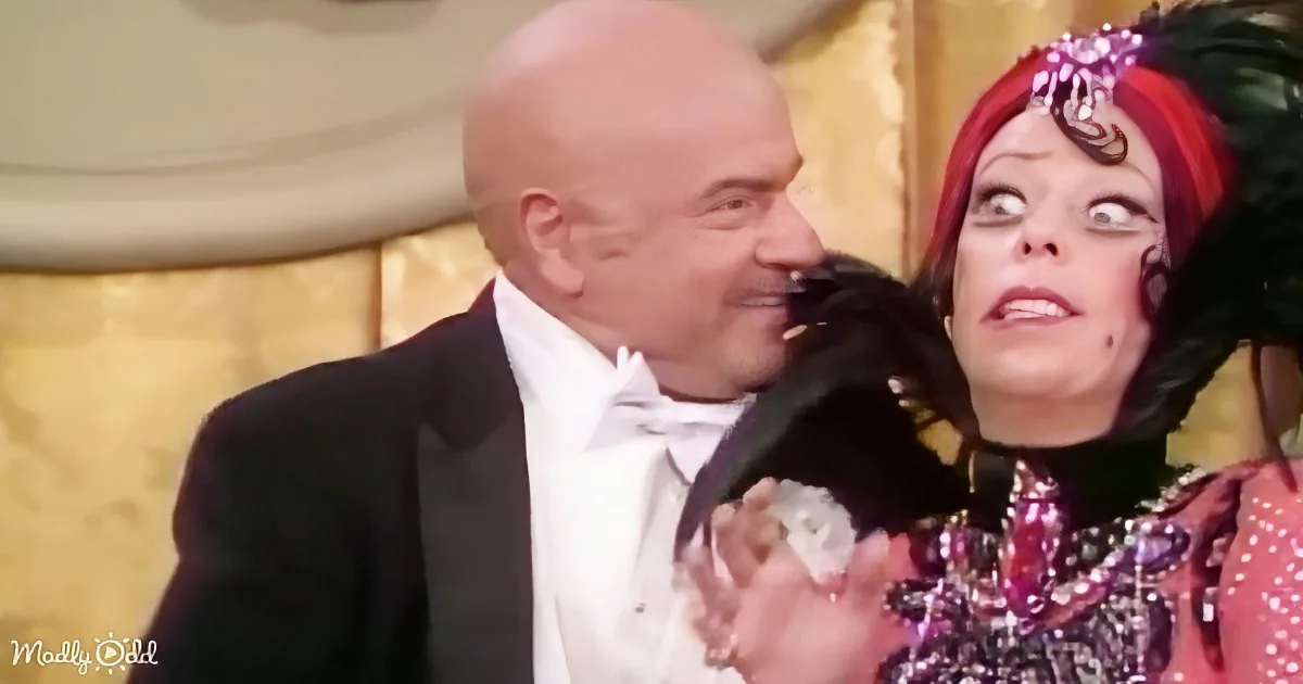 A man in a tuxedo leans in close to a woman in a colorful, ornate outfit, both appearing surprised or shocked.