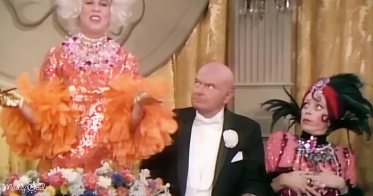 A person in a vibrant, frilly orange dress with a large white wig stands at a table, surrounded by others in formal attire.