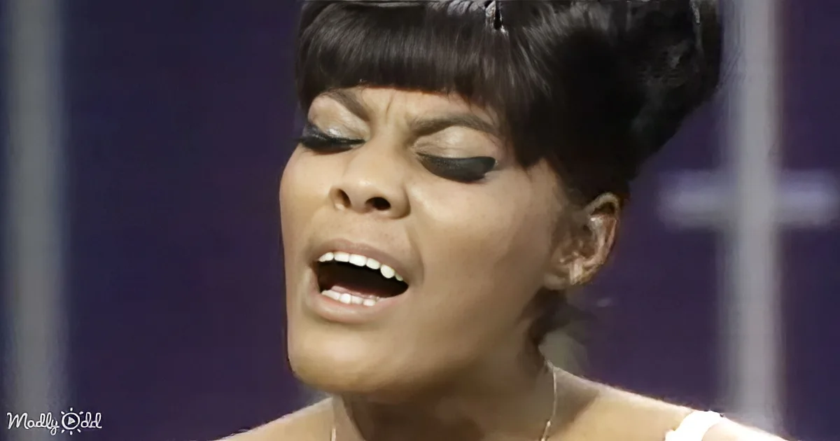 A woman with dark hair styled in a voluminous updo, wearing makeup, and a necklace, appears to be singing or speaking...