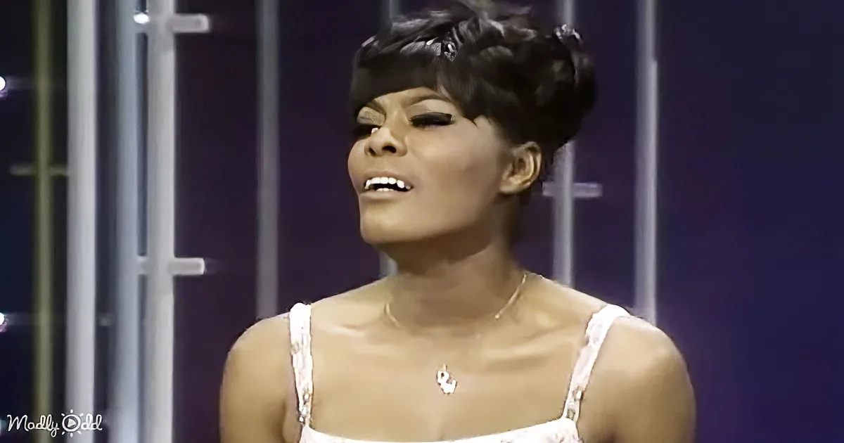 A woman with short, styled hair and a white dress is singing on stage, wearing a necklace.