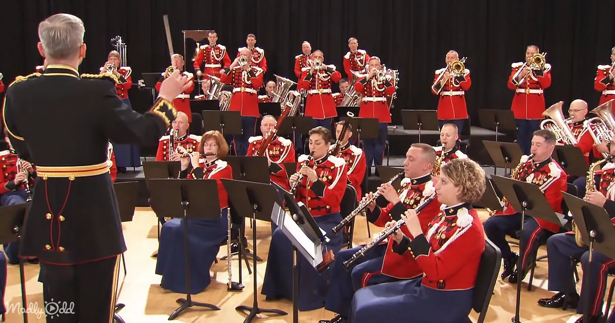 A conductor leads a military band in red uniforms playing various instruments on stage.