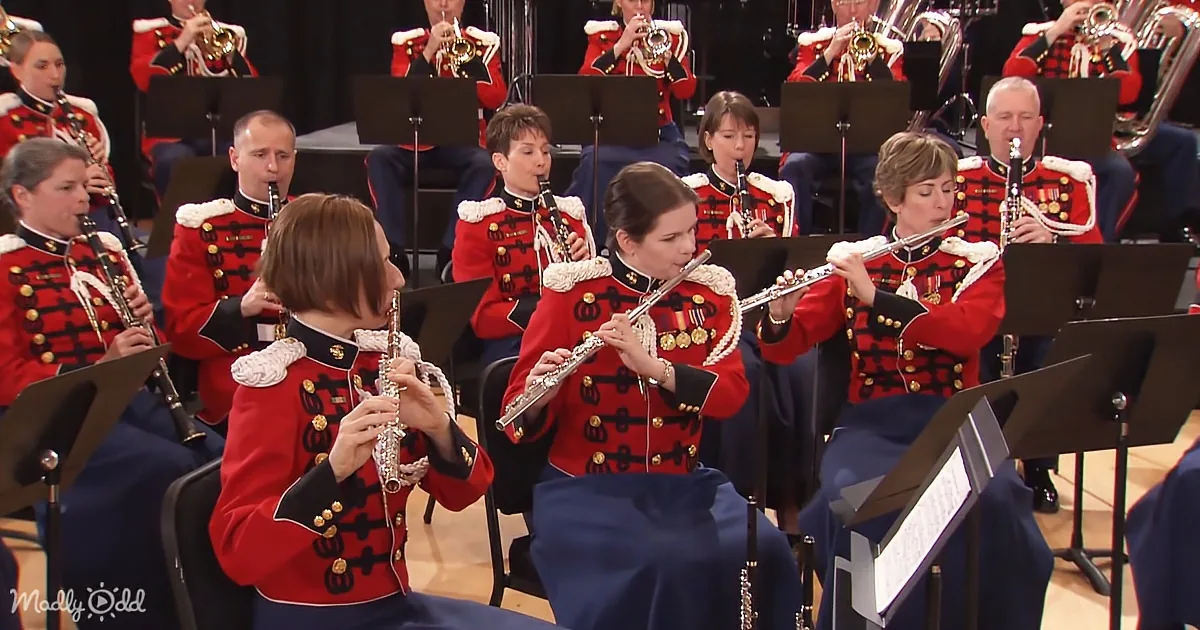 Flutists in red uniforms play in a band, surrounded by other musicians in formal attire.