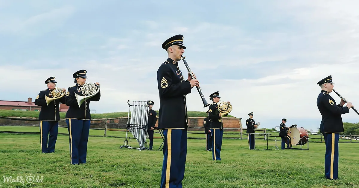 A military band in uniform plays instruments on a grassy field under a cloudy sky.