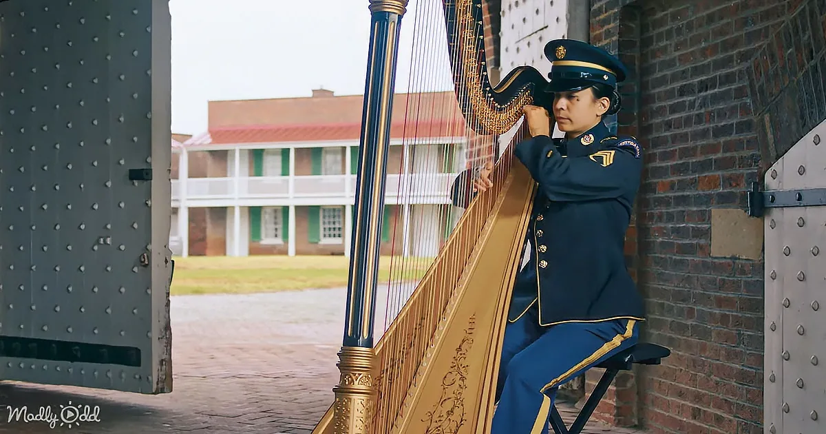 A person in a military uniform plays a golden harp near a brick wall.