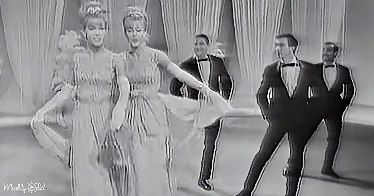 Two women in elegant dresses dance gracefully, surrounded by elegantly dressed men in a formal setting.
