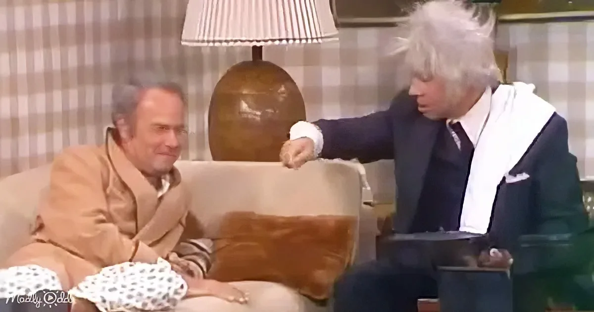 A man in a suit sits on a couch, gesturing with his hand while another man in a tan suit listens.