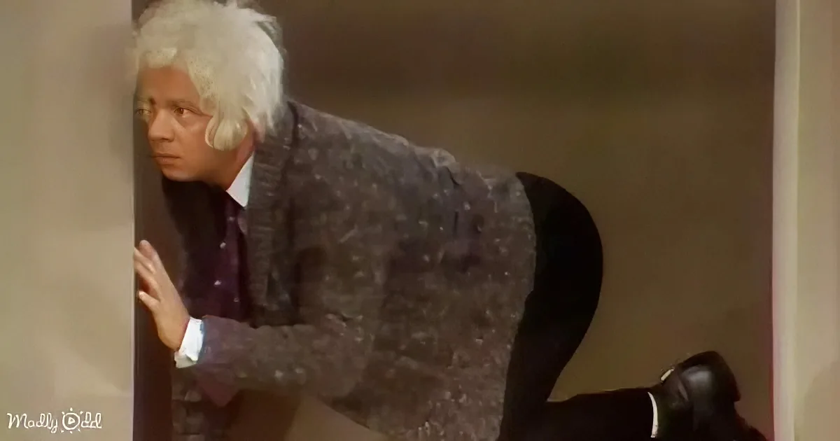 An older man with white hair leans against a wall, wearing a patterned suit and a purple shirt.