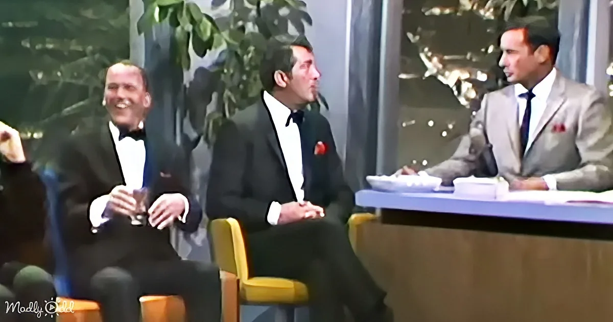 A man in a suit sits at a desk, talking to two men in tuxedos, one laughing.