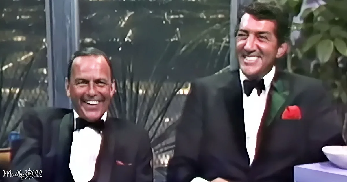 Two men in tuxedos smile, one with a red pocket square, seated at a formal event.