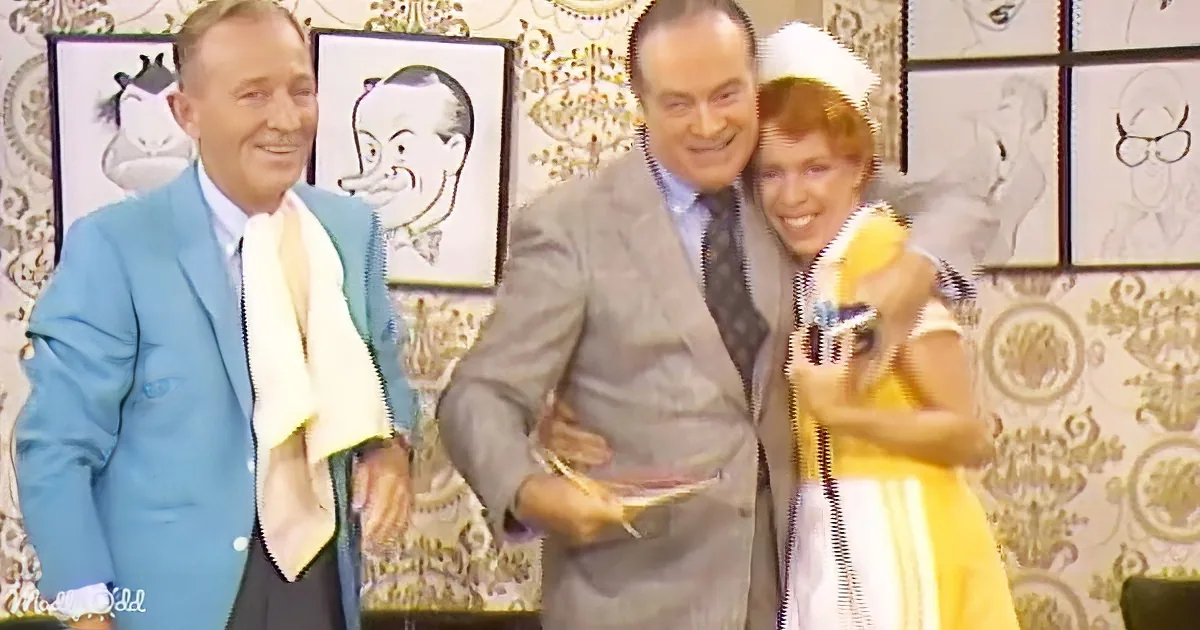 A man in a gray suit holds a book, smiling with two others in a room with patterned wallpaper and caricature portraits.