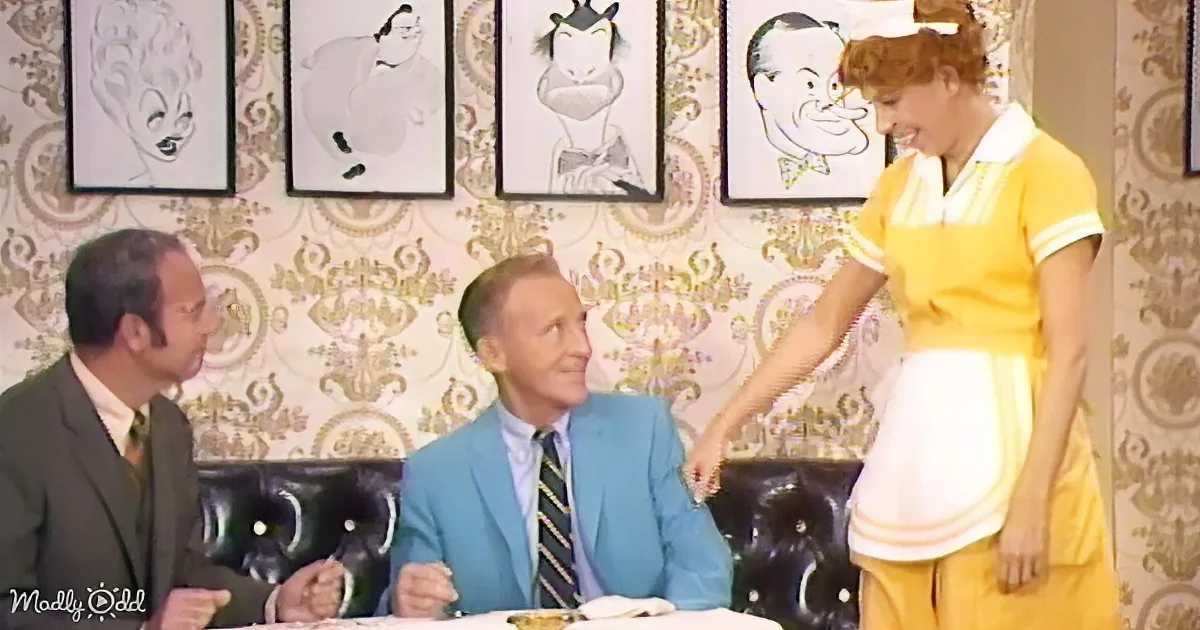 A waitress in a yellow uniform serves a man in a blue suit at a table, while another man in a suit watches.