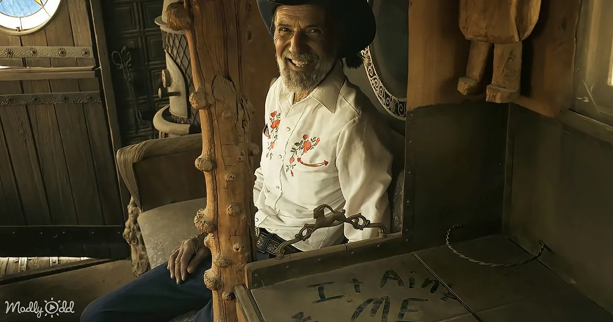 A man in a cowboy hat and white shirt sits inside a rustic wooden structure, smiling.