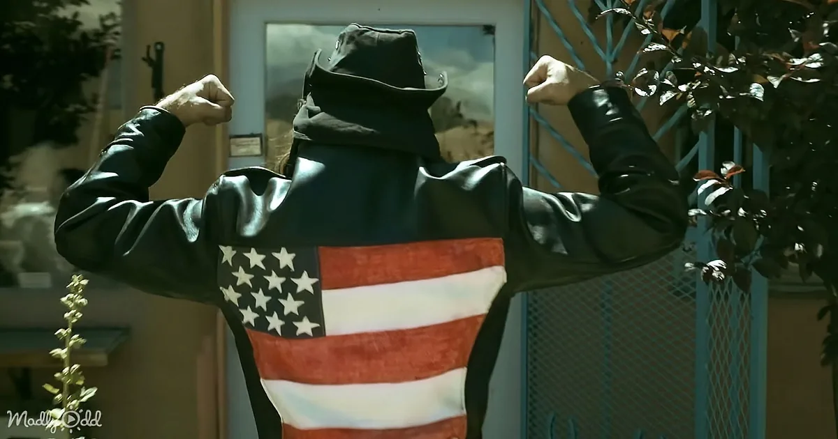 A person in a black leather jacket with an American flag design flexes their muscles in front of a house.
