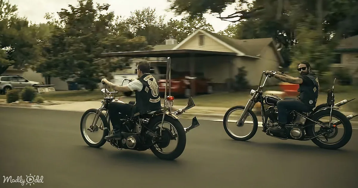 Two motorcyclists in black vests ride side by side on a suburban street, with houses and trees in the background.