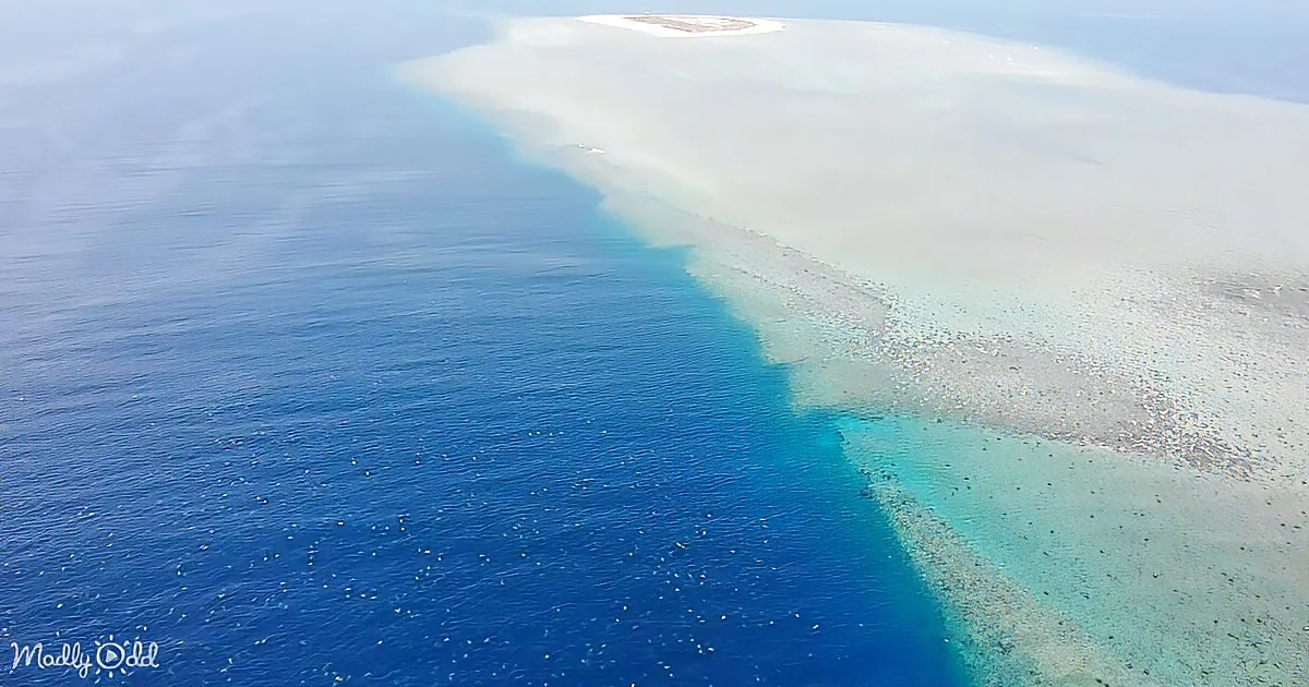 Aerial view of a shallow, turquoise water area transitioning to deeper blue, with a sandy island in the distance.