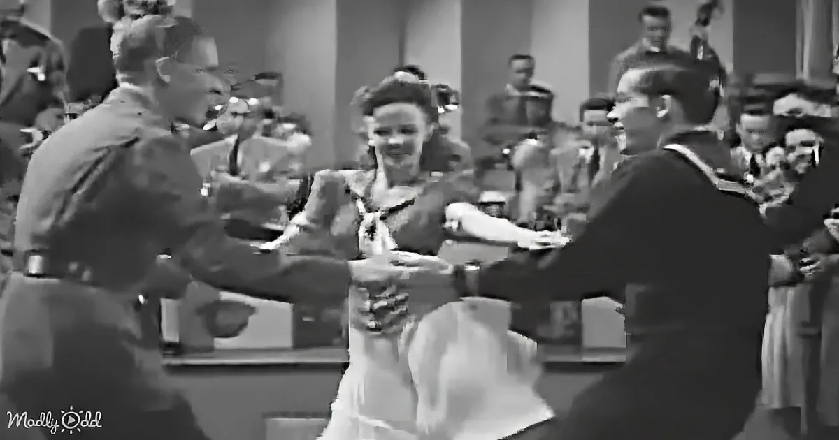 A woman in a dress dances with two men in uniform, surrounded by an audience in a social setting.